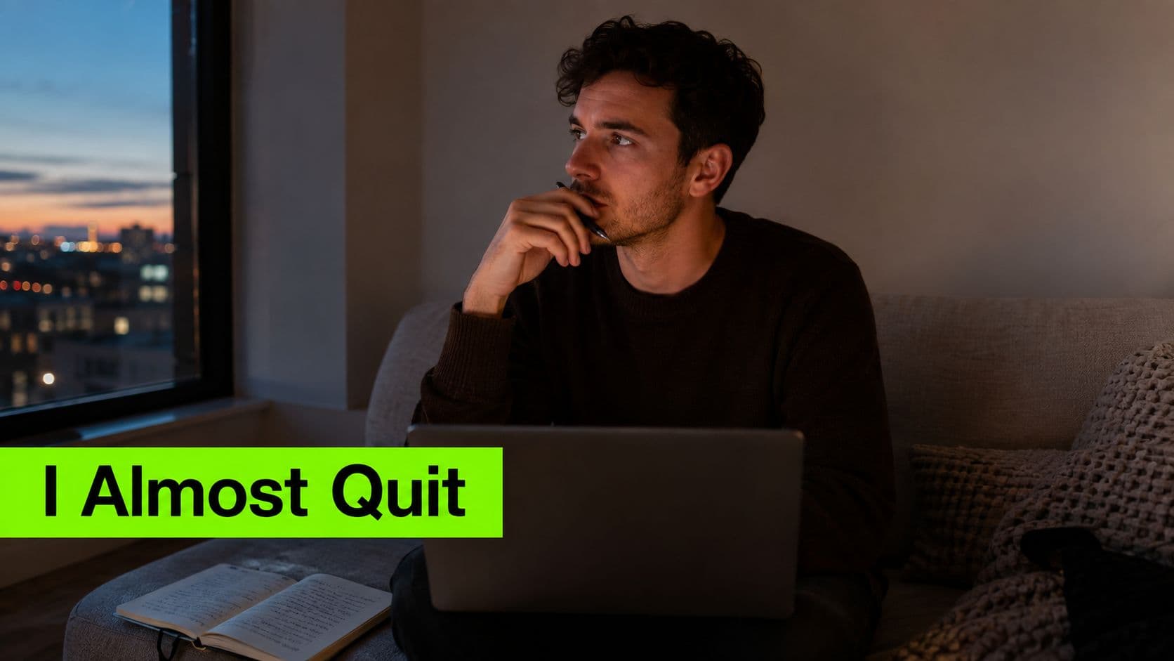 A pensive young man sits on a couch with a laptop and notebook, looking out a window.